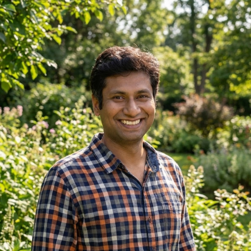 A smiling man in a plaid shirt standing outdoors surrounded by green plants.