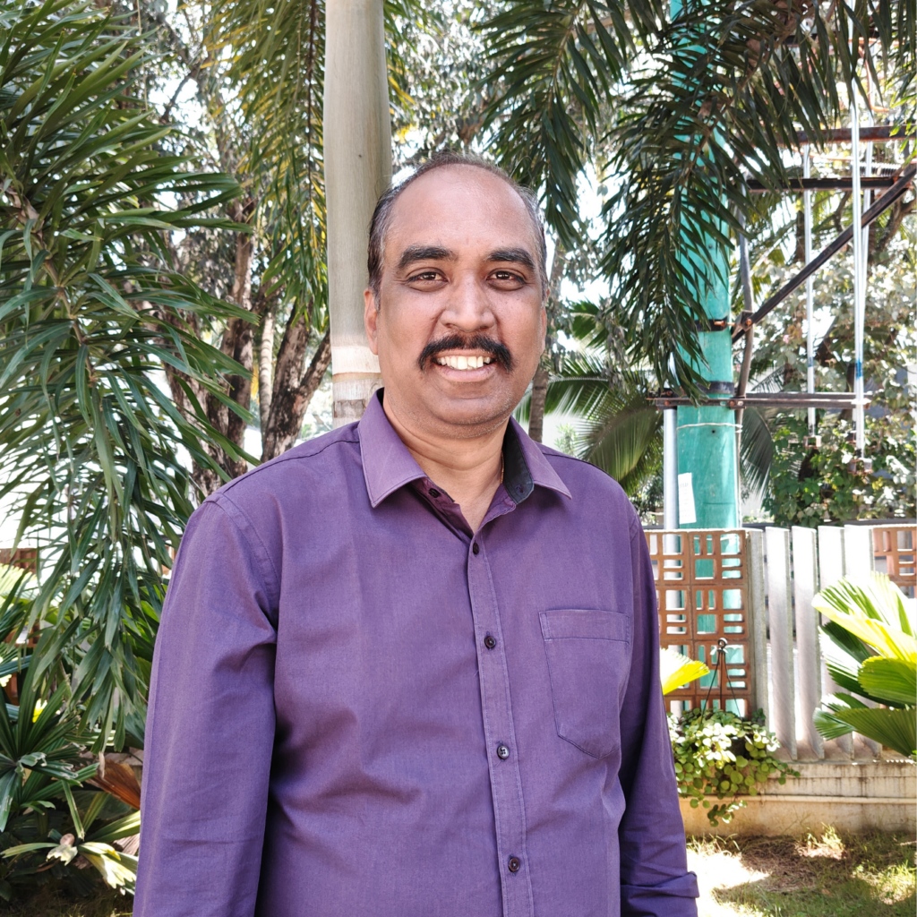 A man with a mustache wearing a purple shirt standing in front of palm trees.