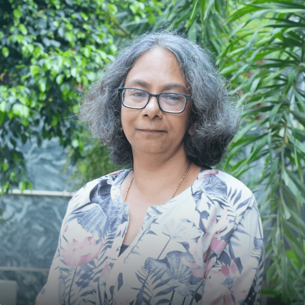 A woman with grey hair and glasses wearing a floral top standing in front of greenery.